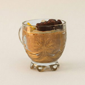 A close up of the Lunis Candle, designed to look like a cup of milk tea. It is poured into a vintage-style clear glass mug with a handle. The wax is a warm, milky brown color, resembling chai, and is topped with a dark brown, biscuit shaped wax embed and yellow floral accents.