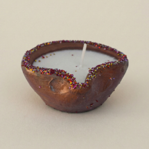 A close-up shot of the Lunis Candle, a small, brown, boat-shaped clay pot candle filled with white wax. The rim of the rustic clay bowl is decorated with colorful purple, gold, and red glitter. The candle features a single white wick and sits against a plain, light beige background.