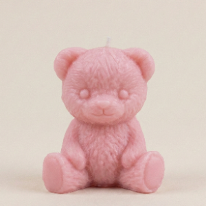 A close up studio shot of a small, solid pink wax candle shaped like a sitting teddy bear. The candle has detailed molded fur texture, small ears, and a white wick protruding from the top of the head. It is sitting on a plain white surface.