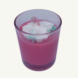 A pink scented candle in a clear glass jar, topped with white floral wax embellishments and a curled pink wick.