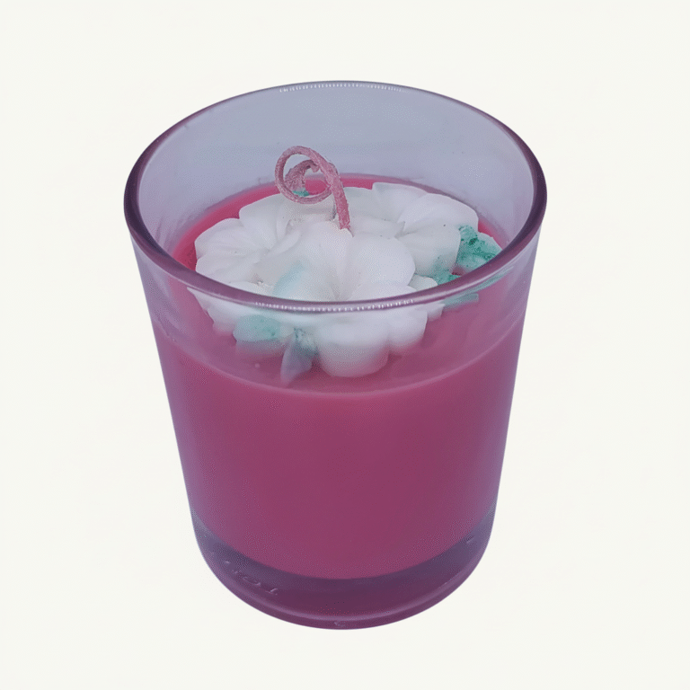 A pink scented candle in a clear glass jar, topped with white floral wax embellishments and a curled pink wick.