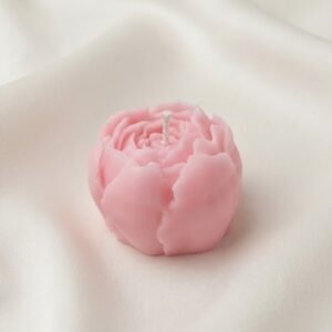 A close up of a pastel pink, peony-shaped decorative candle by Lunis Candles sitting on a seamless white background, featuring detailed wax petals and a central cotton wick.