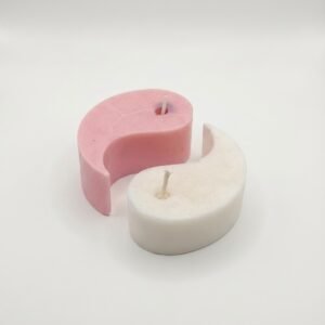 Two interlocking comma shaped pillar candles forming a Yin and Yang symbol. The left candle is a soft pastel pink and the right candle is white. Both have visible wicks and are displayed on a clean white background.