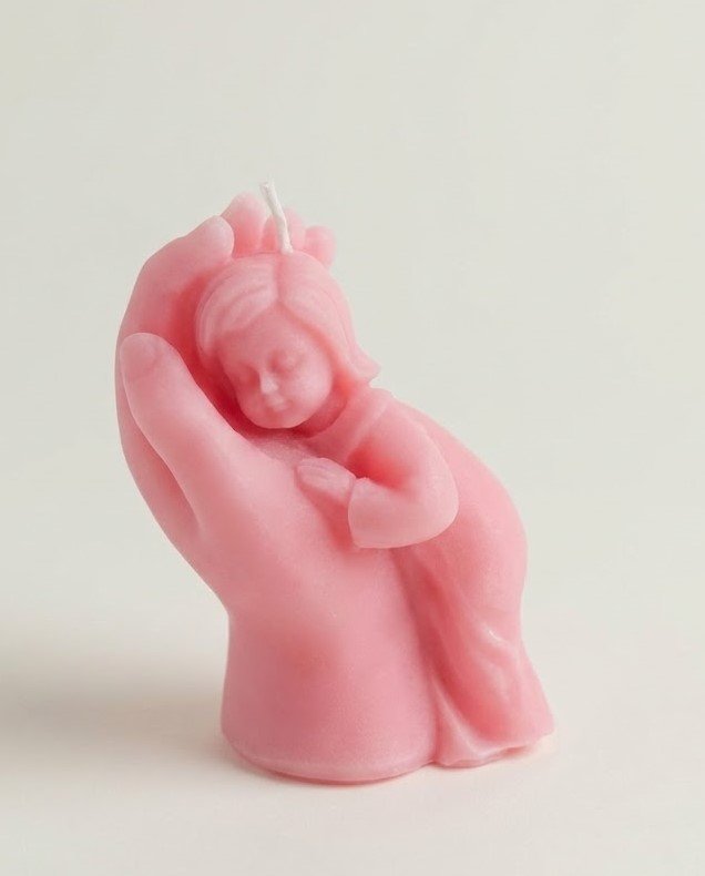 A pink, sculptural wax candle depicting a large hand gently cradling a small, sleeping child, symbolizing protection, comfort, and care against a plain white background.
