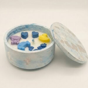 A round, white lidded candle jar made of stone like material featuring pink marble swirls and gold leaf flakes. The matching lid rests against the side of the jar. Inside, the white wax is decorated with small pink wax embeds in the shapes of a teddy bear, baby carriage, rattle, feet, and bottle, along with one small purple heart and one red heart.