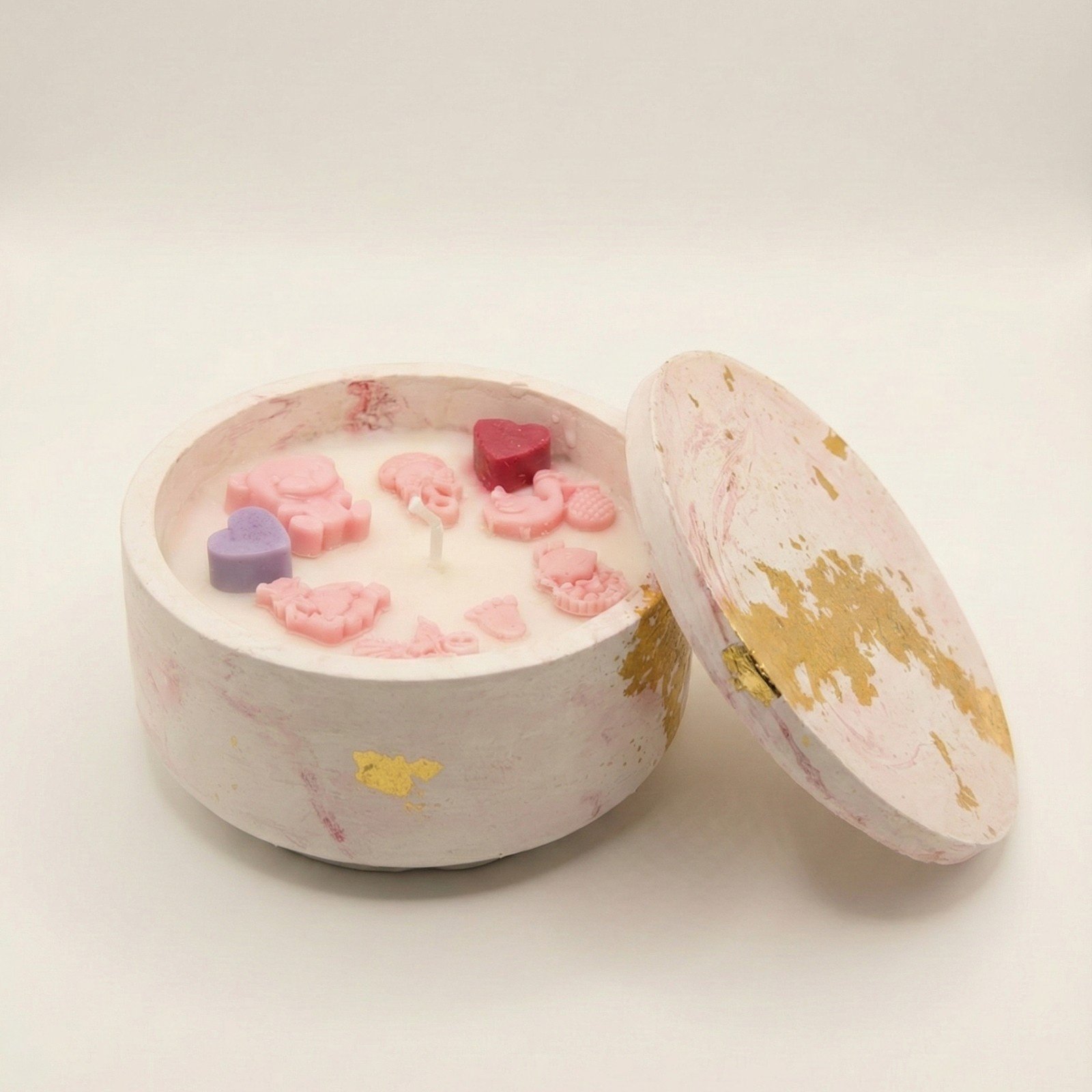 A round, white lidded candle jar made of stone like material featuring pink marble swirls and gold leaf flakes. The matching lid rests against the side of the jar. Inside, the white wax is decorated with small pink wax embeds in the shapes of a teddy bear, baby carriage, rattle, feet, and bottle, along with one small purple heart and one red heart.