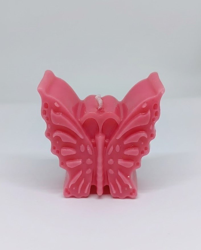 A close up of a bright pink, butterfly-shaped decorative candle standing upright on a white background. The candle features detailed molded wings with small circular patterns and ridges, and a white wick at the top center.