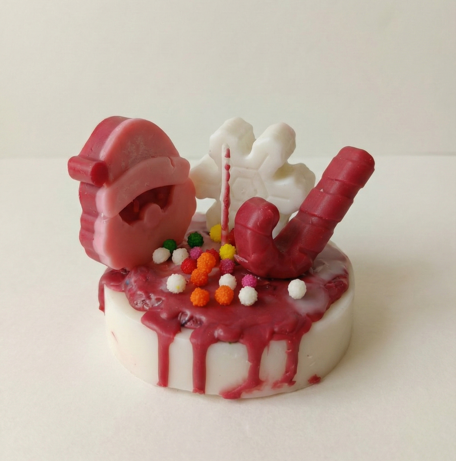 A round, white pillar candle designed to look like a small cake with dripping dark pink wax icing. The top is decorated with three wax figures: a pink and red Santa Claus face, a white snowflake, and a red candy cane. Small, colorful round sprinkles in orange, yellow, and white are scattered across the top. A single wick stands in the center against a plain white background.