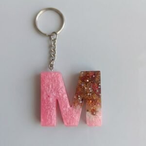A handmade resin keychain in the shape of the letter M. The design features an ombre effect fading from a shimmery pearlescent pink on the left to clear resin packed with chunky gold and copper glitter on the right. It is attached to a standard silver metal keyring and chain.