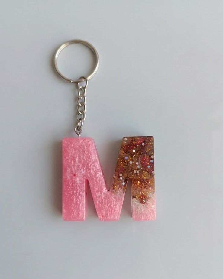 A handmade resin keychain in the shape of the letter M. The design features an ombre effect fading from a shimmery pearlescent pink on the left to clear resin packed with chunky gold and copper glitter on the right. It is attached to a standard silver metal keyring and chain.
