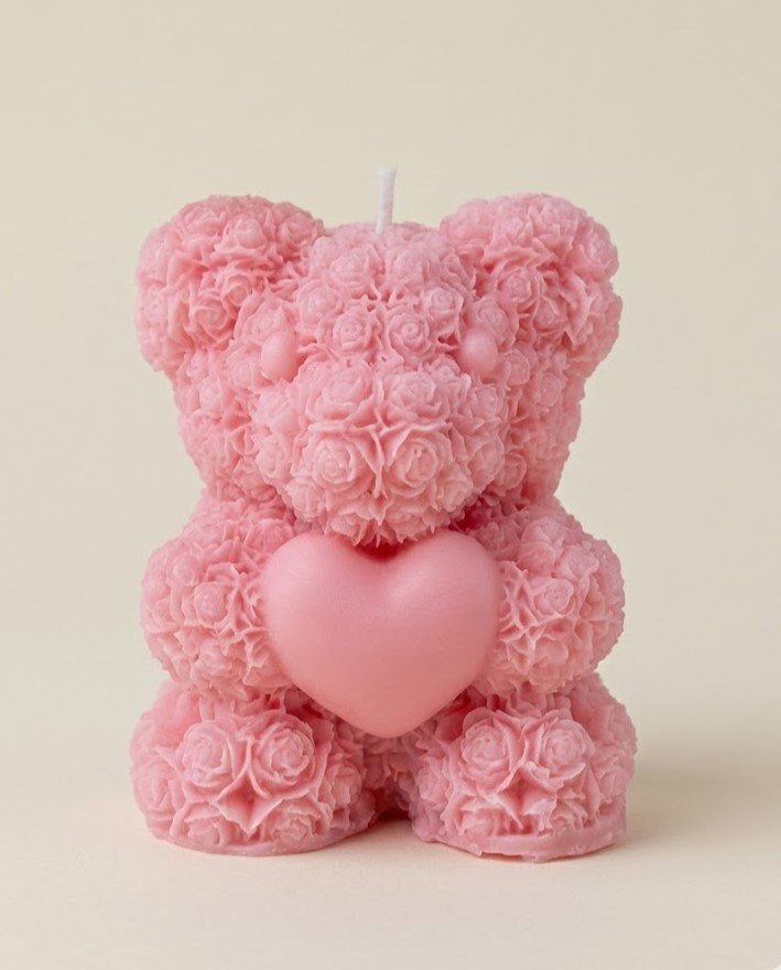 A bright pink, highly textured scented candle shaped like a teddy bear sitting down and holding a smooth heart. The bear's surface is detailed with tiny, densely packed rose shapes.