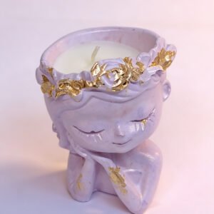 A white scented candle in a decorative holder shaped like a kneeling figure with closed eyes, wearing a floral wreath accented with gold leaf. The figure has a shimmery, pearlescent finish.