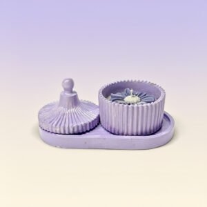 : A small, oval, lilac colored tray holds a matching two piece container. The lid, which has a fluted, round handle and a radiating pleated design, is resting on the left side of the tray. The container base, which has a vertical ribbed texture around its circumference, is on the right and holds a decorative, lilac colored, flower shaped scented candle with a white wick.