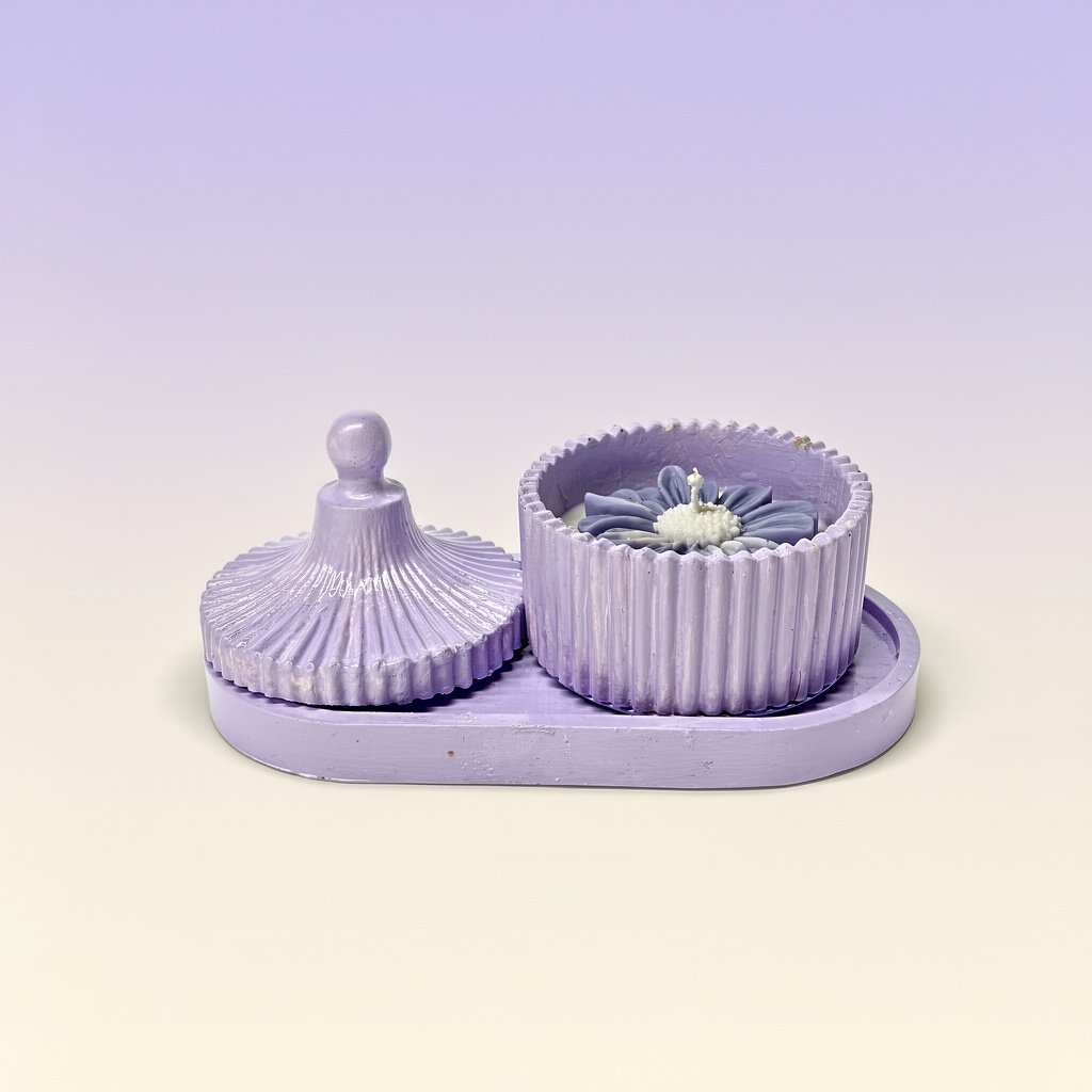 : A small, oval, lilac colored tray holds a matching two piece container. The lid, which has a fluted, round handle and a radiating pleated design, is resting on the left side of the tray. The container base, which has a vertical ribbed texture around its circumference, is on the right and holds a decorative, lilac colored, flower shaped scented candle with a white wick.
