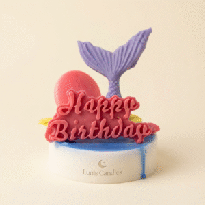 A small, round, scented candle. The base is white with a layer of bright blue wax on top that drips slightly down the side. Topper decorations, likely made of colored wax, include a purple mermaid tail, a hot pink spherical object (like a balloon or pearl), and a large, dark pink cutout text reading