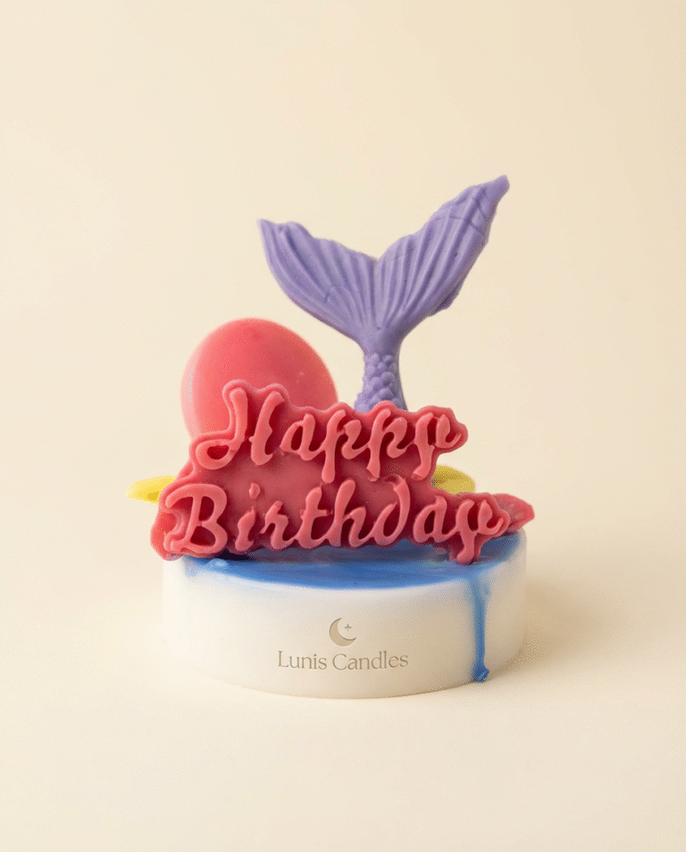 A small, round, scented candle. The base is white with a layer of bright blue wax on top that drips slightly down the side. Topper decorations, likely made of colored wax, include a purple mermaid tail, a hot pink spherical object (like a balloon or pearl), and a large, dark pink cutout text reading "Happy Birthday.