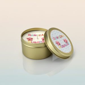 A white, circular scented candle in a gold tin with the lid placed next to it. The candle top features the words 'RAMAZAN MUBARAK' spelled out in colorful letters, with two small pink star shaped wax embellishments between the words.