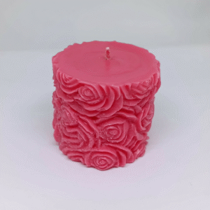 A pink pillar candle with detailed carved rose patterns covering the sides, placed on a light background