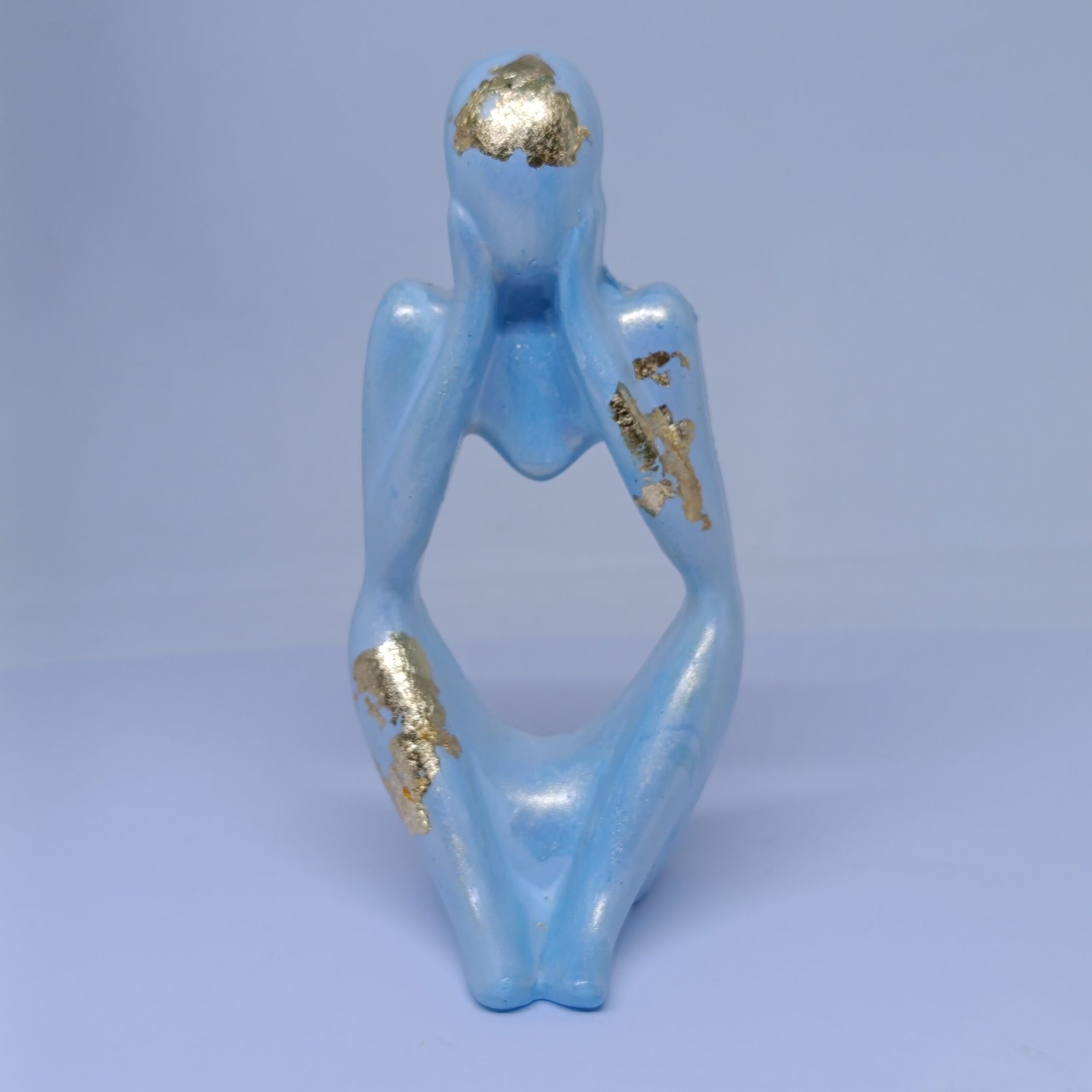 A minimalist blue concrete figurine in a thinking pose with gold foil accents on the head, arm, and leg, displayed against a light background.