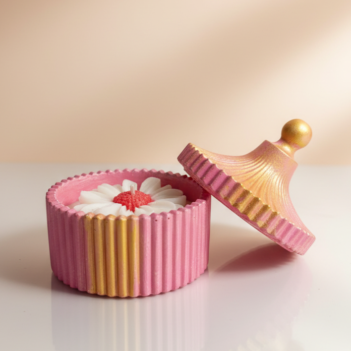 A pink and gold decorative candle jar with a ribbed design, holding a white daisy shaped scented candle inside, with the lid placed beside it.