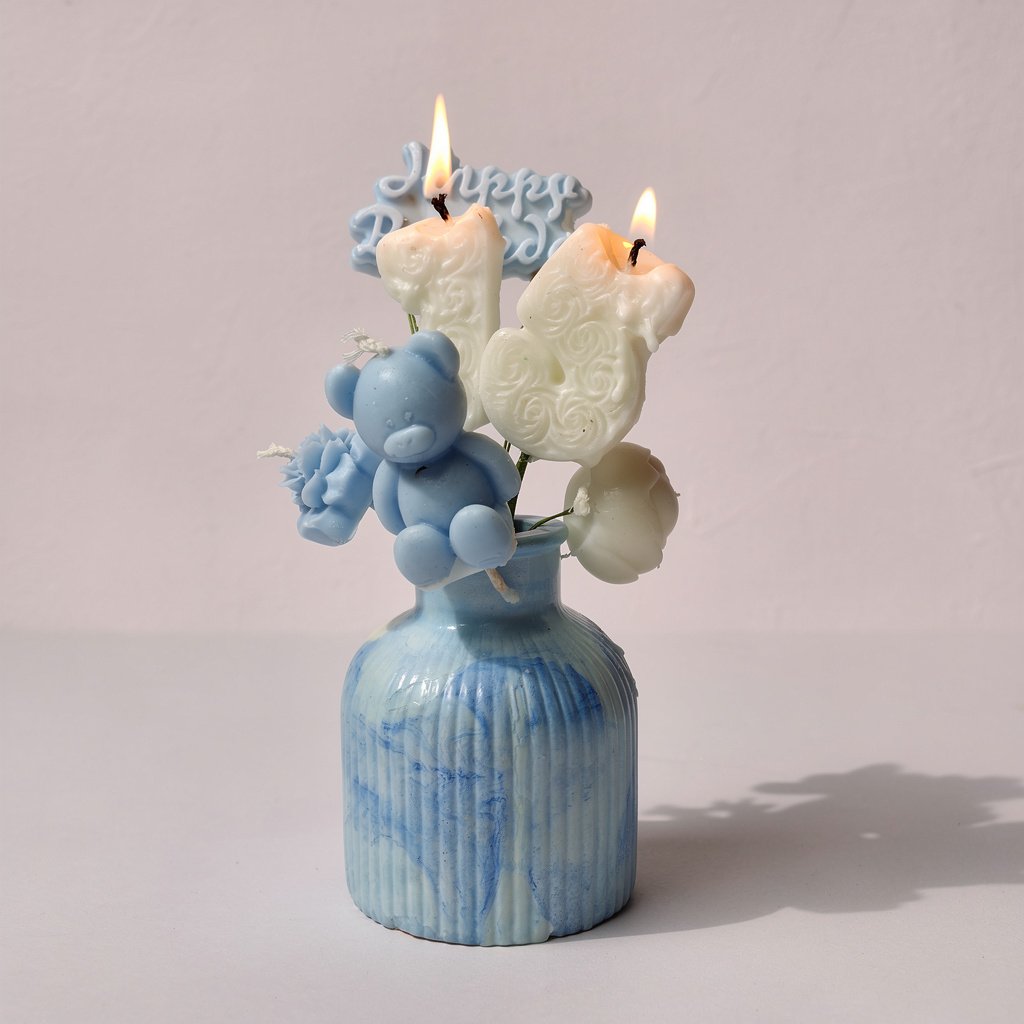 ● A lit, scented candle display in a small blue and white marbled ceramic vase, with a small blue teddy bear and blue floral accents. A faint blue 'Happy Birthday' message is visible behind the flames.