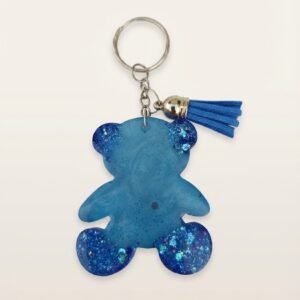 Blue resin teddy bear shaped keychain with a silver keyring, chain, and a small blue faux suede tassel. The resin is a translucent, medium blue color with fine glitter throughout, and larger, chunky iridescent blue glitter is concentrated on the ears and feet