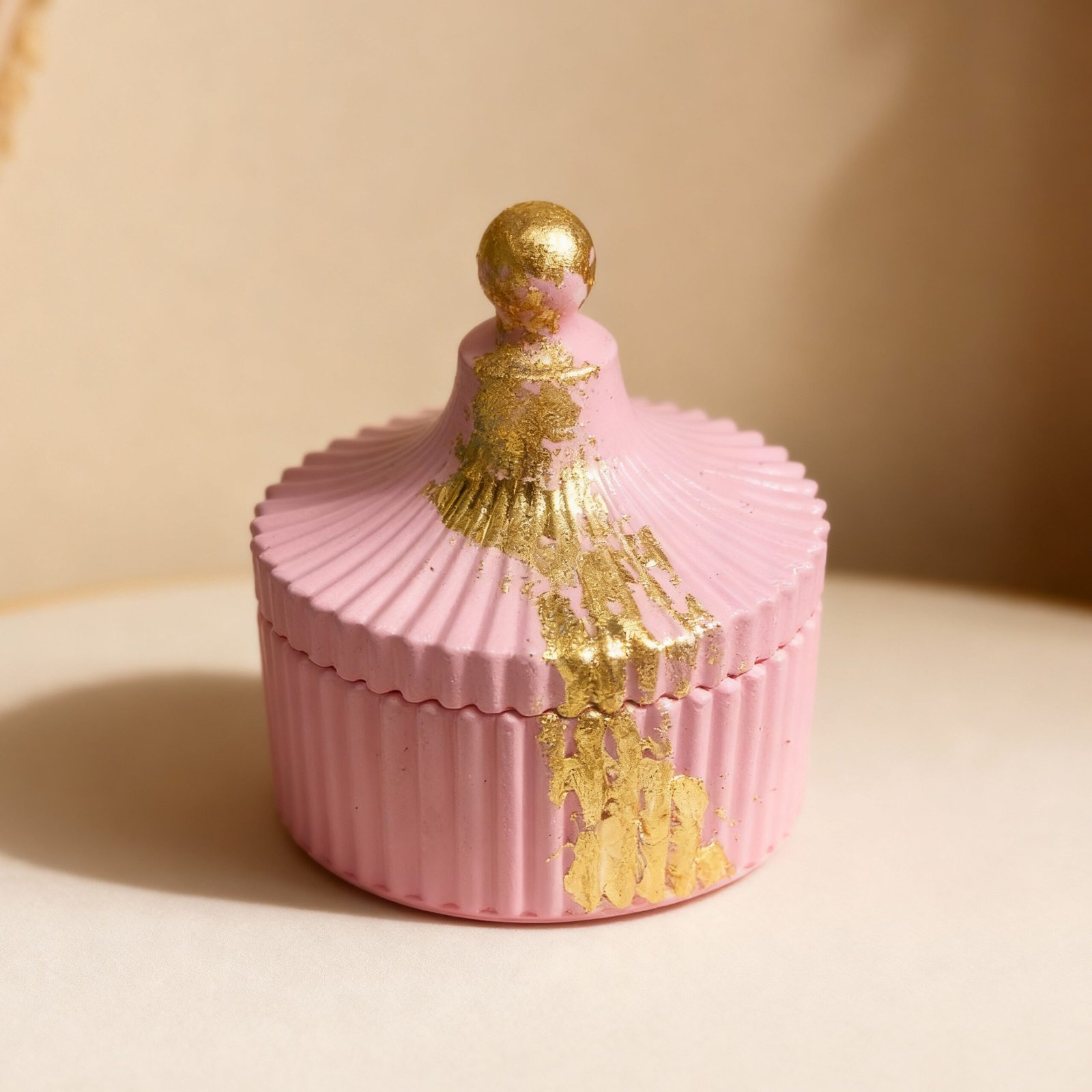 A round fluted concrete trinket box in a rose pink color featuring a lid with a knob and heavy dripping gold leaf detail on the top and side.