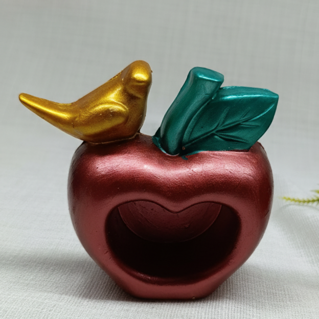 A handcrafted concrete apple shaped trinket dish with a heart shaped hollow center, green leaves, and a golden bird perched on top.