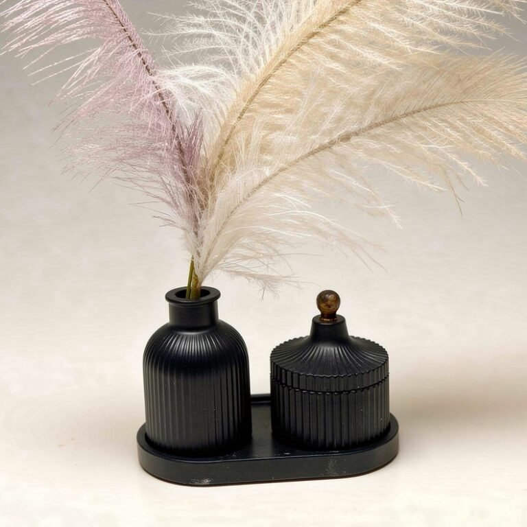 Two black ribbed concrete containers on a shared base, one holding pampas-style stems, displayed against a light fabric backdrop.