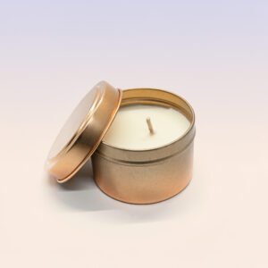A round, bronze colored metal tin candle. The lid is partially resting on the rim of the tin, revealing a smooth, white wax candle inside with a single centered cotton wick. The tin is set against a plain white or light background.