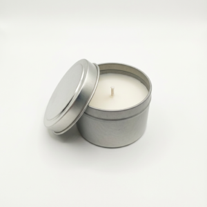 A simple, white, scented soy or wax candle set in a round, silver metal tin. The lid is leaning against the side, showing the centered wick and smooth surface of the candle wax.